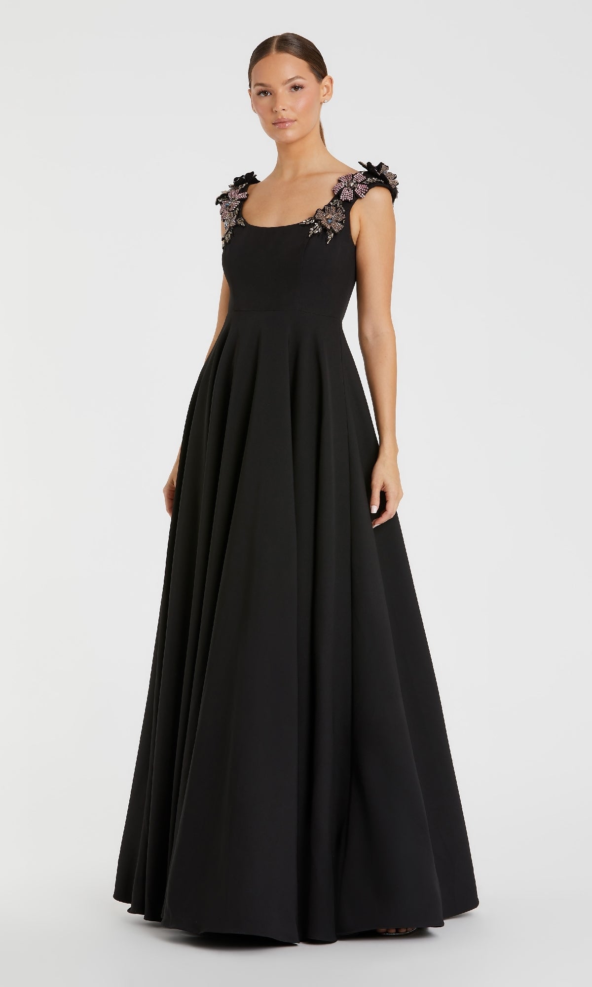 A woman stands against a plain white background wearing the Mac Duggal 11949 Long Black A-Line Formal Dress, featuring a fitted bodice and floral embellishments on the shoulder straps.